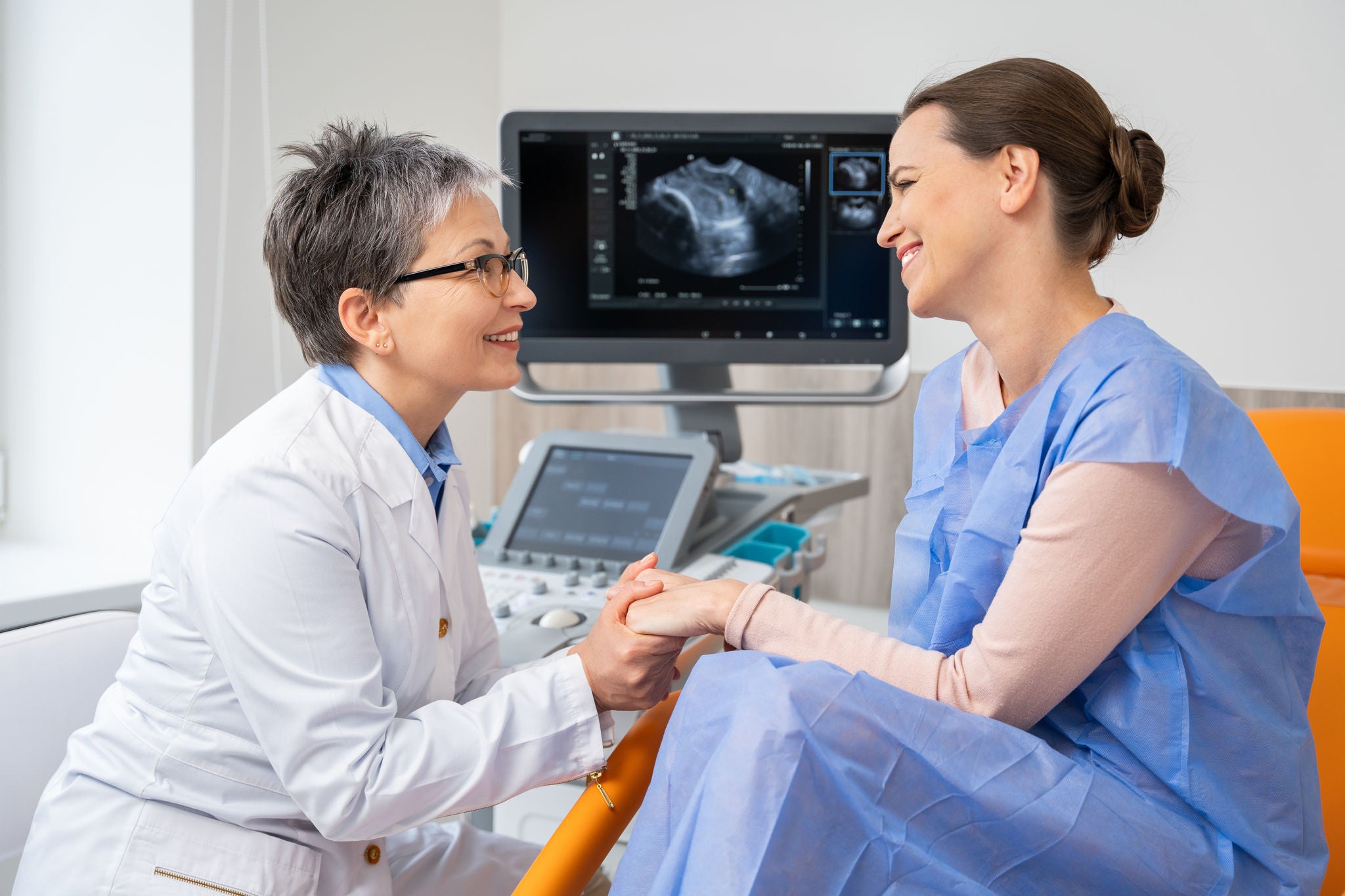 A smiling doctor holds the hand of a happy expectant mother during a maternity ultrasound session, with the sonogram visible in the background. High quality photo
