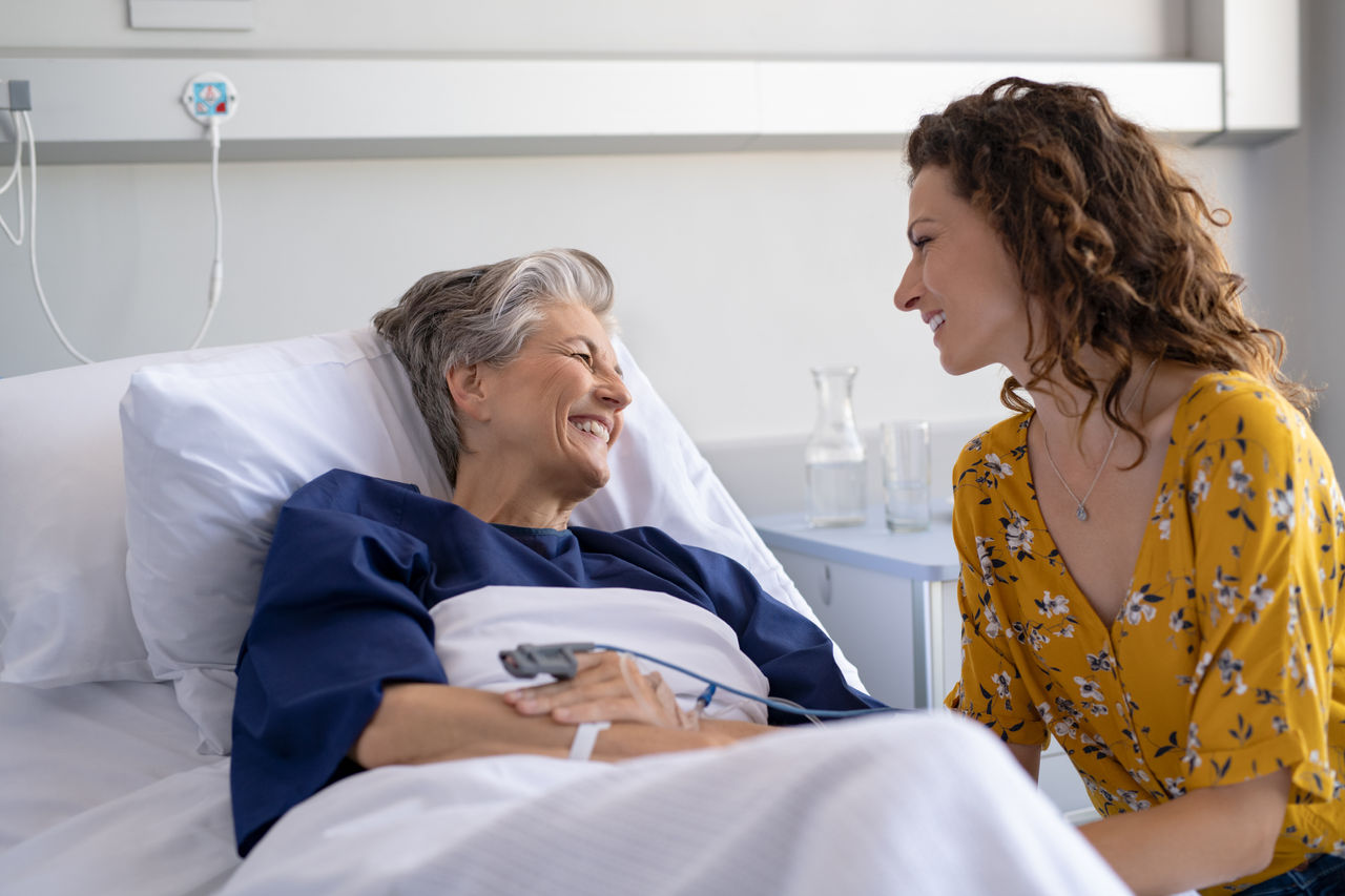 Young daughter visiting sick old mother in hospital. Happy senior patient smiling while in conversation with her lovely daughter. Granddaughter visiting and cheering her hospitalized grandmother lying in bed at hospital ward after surgery.