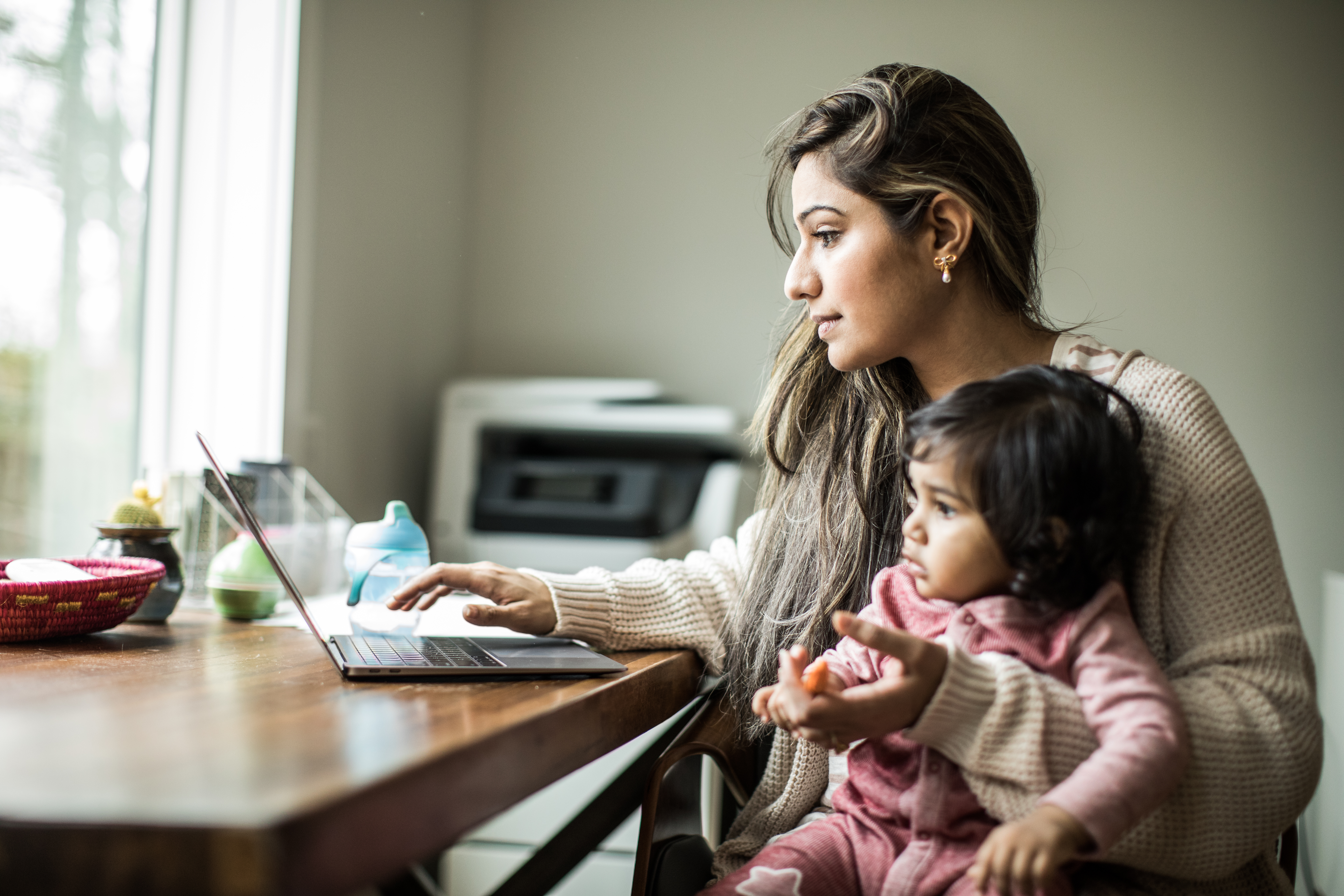 A focused mother holds her baby on her lap while working on a laptop at a wooden table near a window. The scene depicts the challenges and reality of balancing work and childcare at home.
