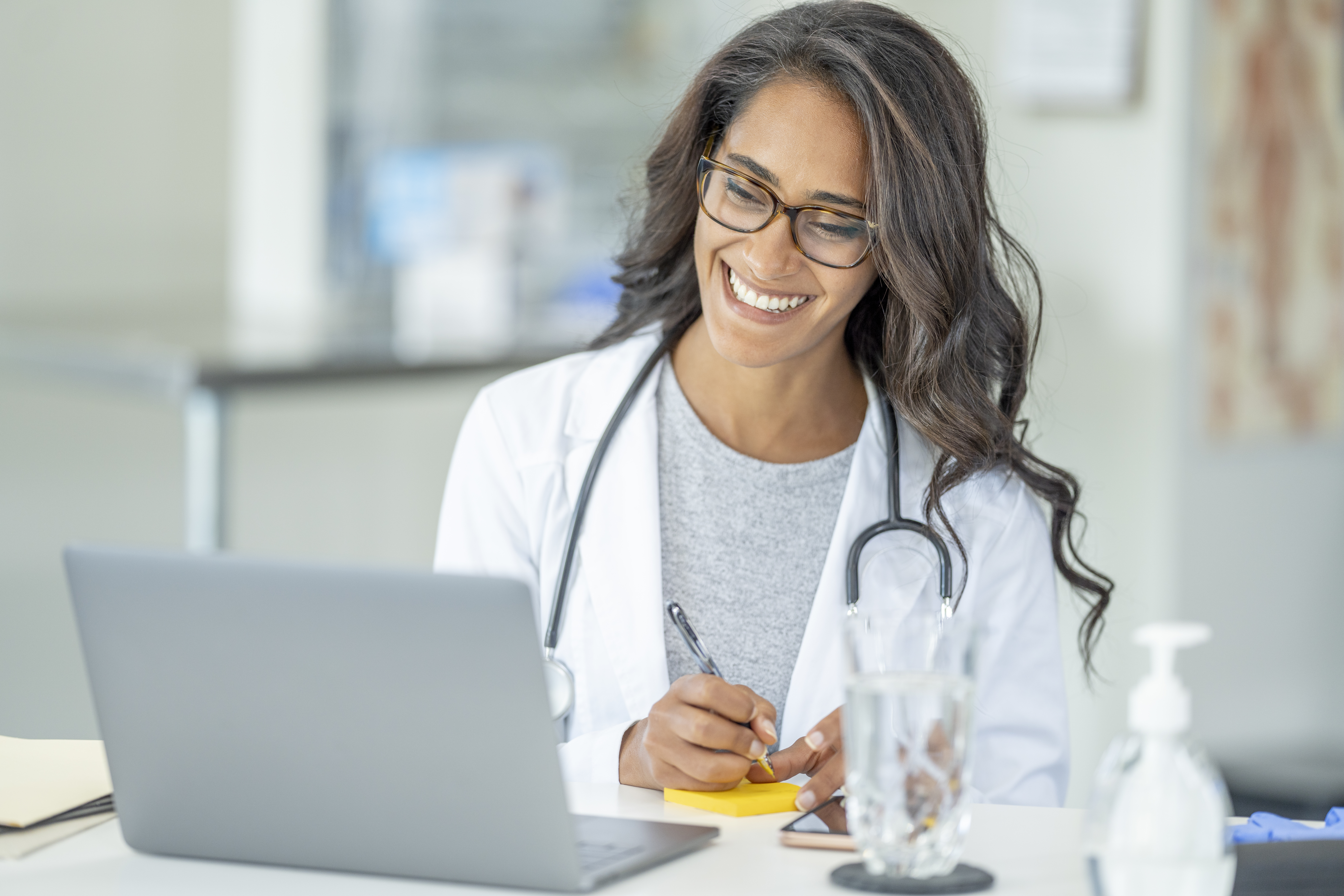 A smiling female doctor with glasses and a stethoscope is seated at a desk. She is writing on a sticky note and looking at a laptop, suggesting a telehealth consultation or office work.