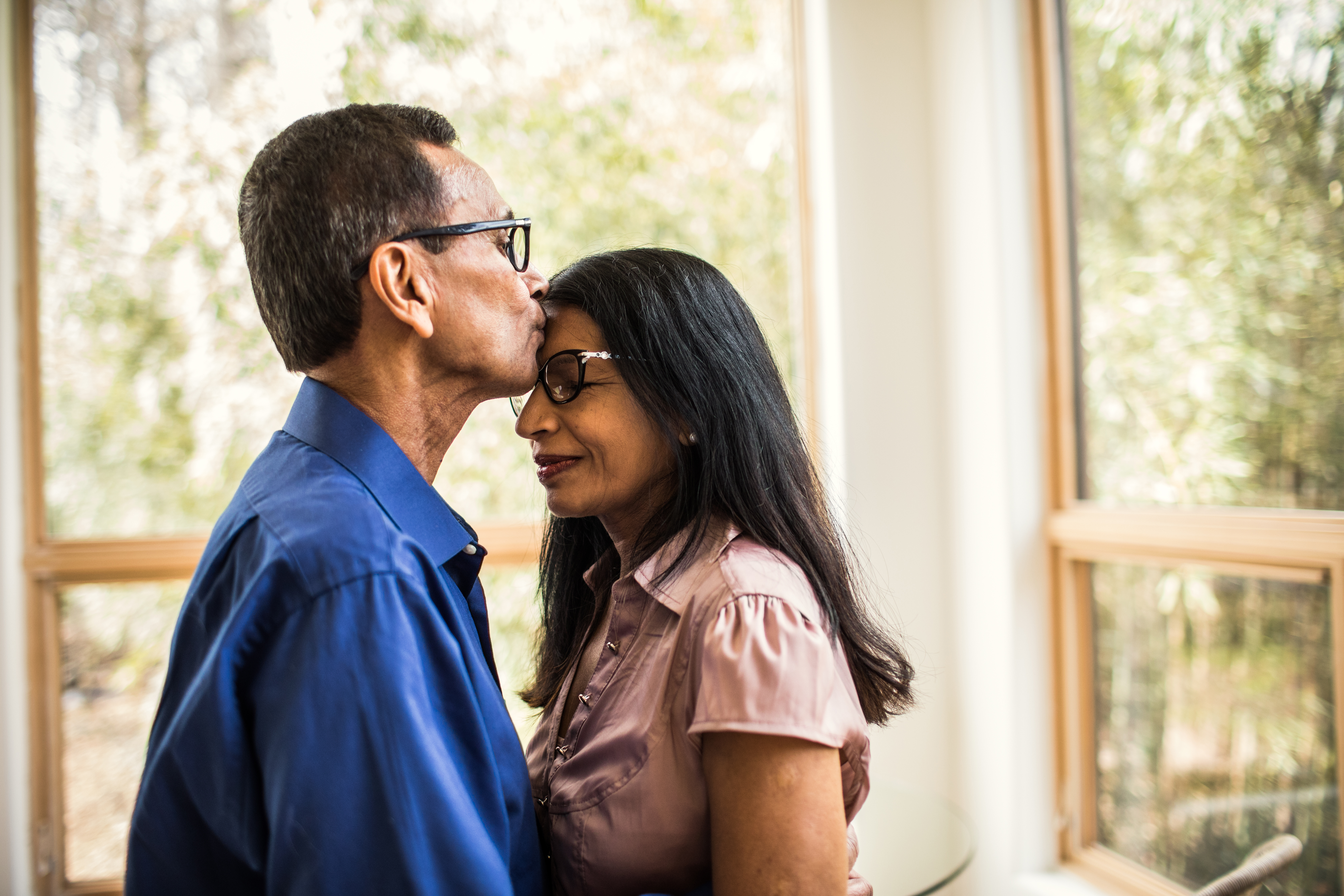 An Indian man with glasses is tenderly kissing the forehead of an Indian woman with glasses, who has her eyes closed, implying a moment of deep affection. Natural light filters through the large windows behind them.