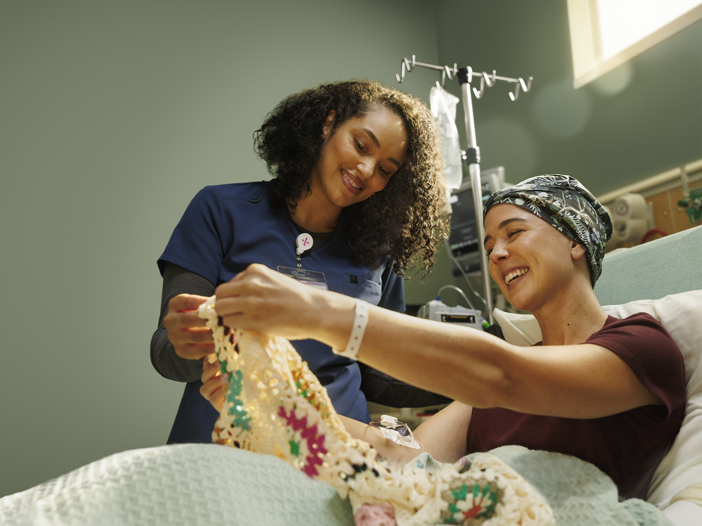 A patient receiving chemo therapy shows their knitting to a Nurse. Both have smiles on their face showing humankindness