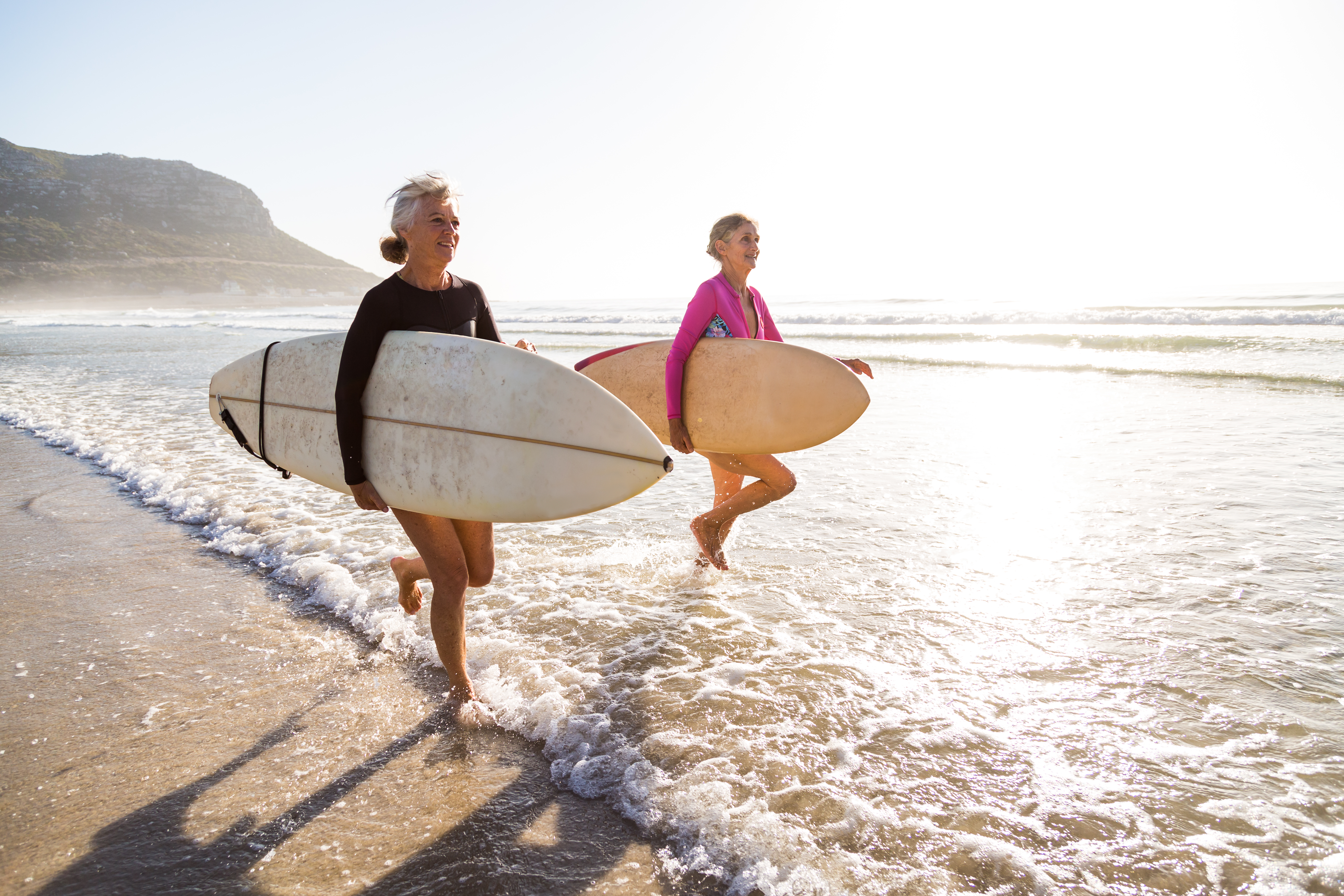 Two smiling elderly women, carrying surfboards, run joyfully into the ocean. The sunny beach scene highlights active aging and pursuing hobbies later in life.